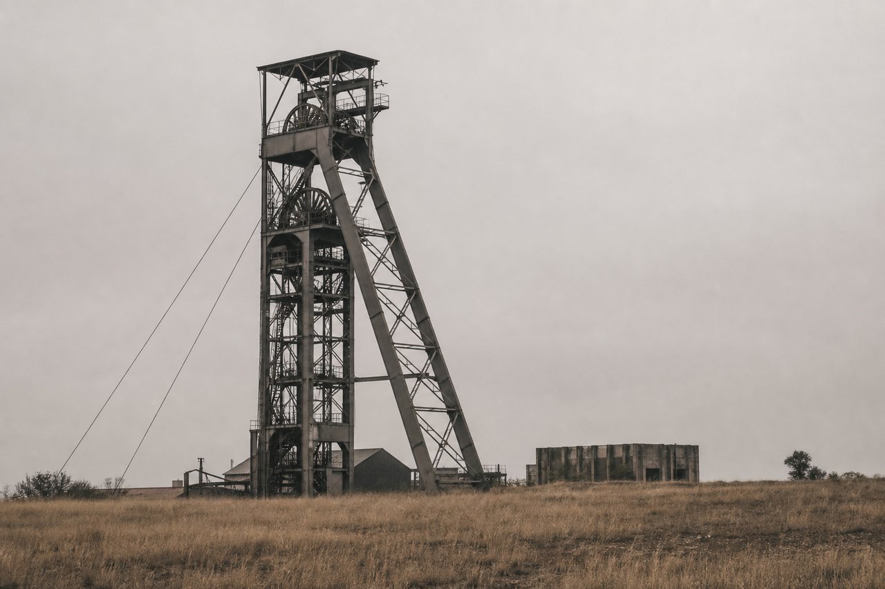 The steel headframe of a deep South African mine shaft against a flat overcast sky, dry pale grassland at its foot